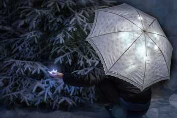 Girl squats with outstretched arm with lights in the woods by the Christmas tree with a luminous umbrella.