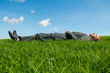 Relaxed businessman lying on his back on a sunny patch of bright green grass in a field with blue...