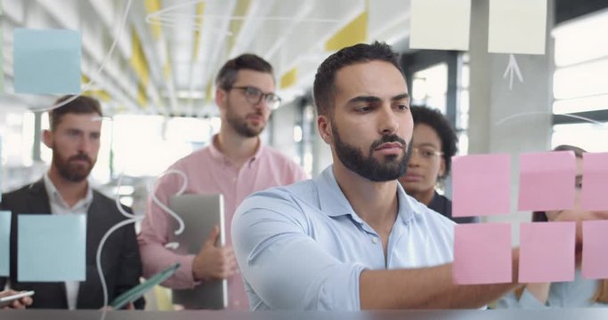 Close up of male office worker thinking and using glass board ato solve problem task. Diverse coworkers at meeting standing and looking at team leader near glass wall with sticky notes.