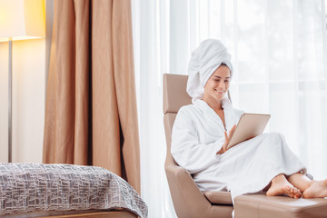 Good looking young American European pretty woman in white bathrobe and towel looking at home goods with her high-speed internet tablet and online store sitting in cozy bedroom - obrazy, fototapety, plakaty