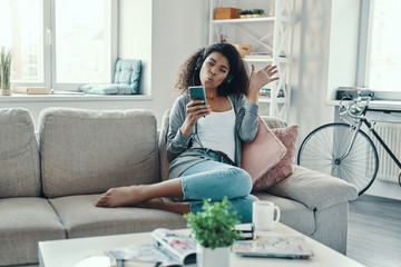 Playful young African woman in casual clothing listening music and making a face while resting at home