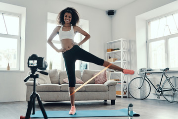 Obraz na płótnie Canvas Full length of attractive young African woman exercising using strap and smiling while making social media video