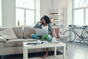 Beautiful young African woman in casual clothing using laptop while resting at home