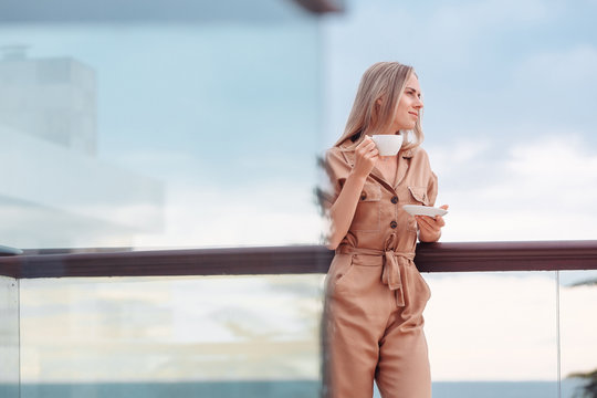 Portrait of a happy smiling caucasian woman in stylish overalls with a cup of coffee meeting the day standing on the hotel terrace with a gorgeous view of the sea and nature - Powered by Adobe