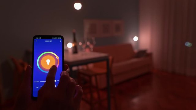 Smart home concept. Woman turning on light in room and changing color with her smartphone and smart house app for cozy relaxing evening. UHD, 4K