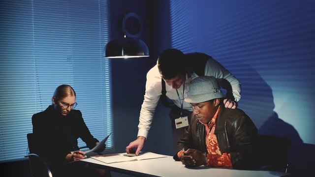 Detective uses force in questioning the accused black man while another female detective reading documents of the interrogation.