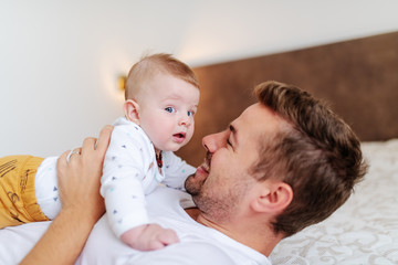 Smiling caucasian handsome dad lying on bed in bedroom and holding his loving adorable six months old son on his chest. Unconditional love concept.