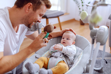 Happy adorable 6 months old baby boy lying in baby rocker chair and looking at toy that his caring dad holds. Healthy upbringing concept.