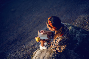 Top view of attractive caucasian brunette in floral dress and with headband sitting on the rock on shore, eating apple and writing diary. - obrazy, fototapety, plakaty