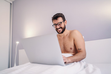 Young shirtless smiling caucasian bearded tattooed man sitting on bed in bedroom and using laptop.