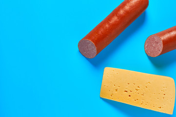 One piece of cheese in form of triangle near two halves of long sausage lies on blue desk on kitchen or market. Space for text. Top view