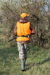 A man with a gun in his hands and an orange vest on a pheasant hunt in a wooded area in cloudy weather. Hunter with dogs in search of game.