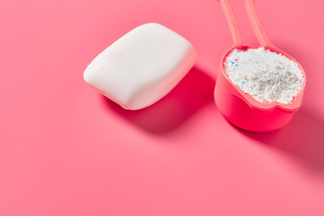 Plastic measuring spoon full of washing powder for cleaning clothes near antibacterial soap lies on pink countertop in laundry. Space for text