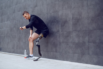 Full length of handsome caucasian sportsman with artificial leg leaning on wall, looking at wristwatch and resting from running. Next to him is bottle with refreshment.