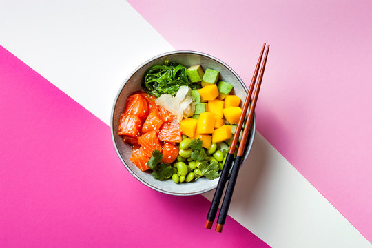 Hawaiian salmon poke bowl with seaweed, avocado, edamame, mango and pickled ginger. Top view, bright pink background