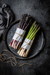 Bunches of fresh green and purple asparagus on vintage metal tray over dark grey rustic background. Top view, copy space