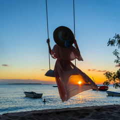 Beautiful girl in a straw hat and pareo swinging on a swing on the beach during sunset of Zanzibar island, Tanzania, Africa. Travel and vacation concept - obrazy, fototapety, plakaty