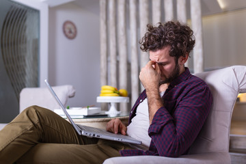 Young tired man sitting at home surrounded with papers and laptop computer having headache holding his hand on head having pain closing his eyes being sleepy and exhausted. Tiredness