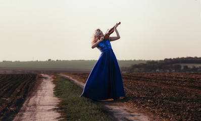 woman in long dress playing violin on background of field path with a club of dust, girl engaged in musical art, performance on nature, concept passion in music © fantom_rd