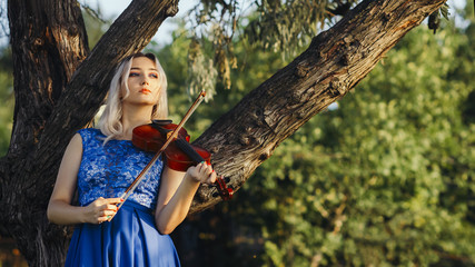 beautiful romantic girl playing violin lean on tree outdoors, young woman in long elegant dress...