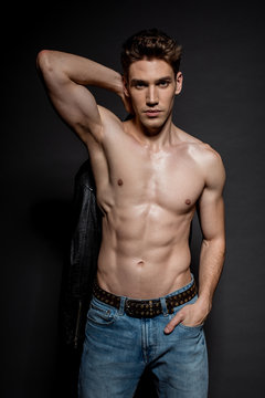 sexy young man with muscular torso and biker jacket posing with hand in pocket on black background