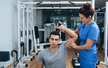 Mixed races athlete with a shoulder injury in a rehabilitation center. Physiotherapist helps...