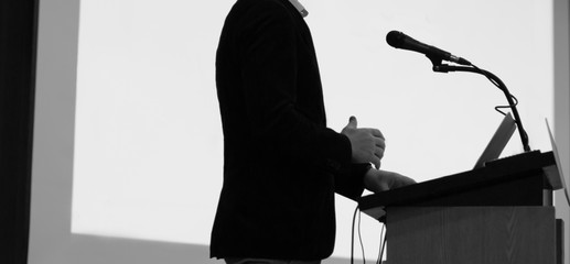 Speaker giving a talk at a corporate business conference. Audience in hall with presenter in front of presentation screen. Corporate executive giving speech during business and entrepreneur seminar. 
