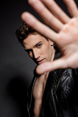 selective focus of sexy young man with muscular torso in biker jacket showing hand at camera on black background