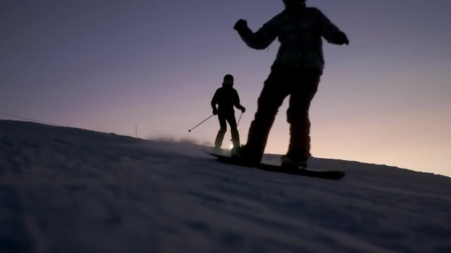 Slowmotion footage of a skier and snowboarder riding the slope during a beautiful sunset.
