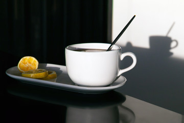 a pottery white cup on a black table and its shadow from the bright sun; tea break with lemon