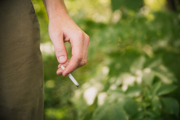 Young man in a moody dark woods forest area holding a roll up tobacco cigarette in his hand resting against leg burning away dropping ash on the floor and having the risk of cancer QUIT