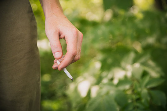 Young man in a moody dark woods forest area holding a roll up tobacco cigarette in his hand resting against leg burning away dropping ash on the floor and having the risk of cancer QUIT - Powered by Adobe