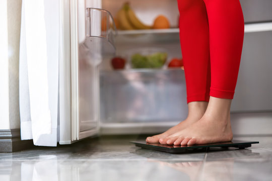 woman's legs standing on scales to measure weight in the kitchen near open fridge full of fruits and vegetables, dietology and nutrition