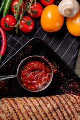Top view bowl of chili, tomato and garlic sauce on black stone background. Tasty barbecue ketchup. Red bbq sauce, grilled meat and vegetables on rustic background.