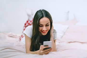 Attractive young smiling woman lying in bed in cozy light colored bedroom texting message on her mobile phone