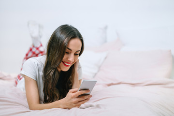 Attractive young smiling woman lying in bed in cozy light colored bedroom texting message on her mobile phone