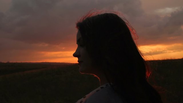 Silhouette of a girl with long hair on the setting sun in a field, a strong wind blows up the girl's hair. Close-up. Slow motion.