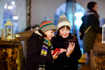 Obraz premium Two little kids, boy and girl having fun on traditional Christmas market during strong snowfall with taking photos with mobile smartphone or phone. Happy children, siblings and best friends