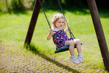 Cute adorable toddler girl swinging on outdoor playground. Happy smiling baby child sitting in chain swing. Active baby on sunny summer day outside