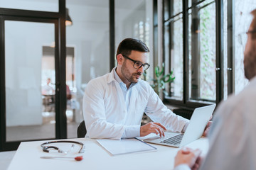 Portrait of a serious doctor looking at laptop while on consultation with a patient.