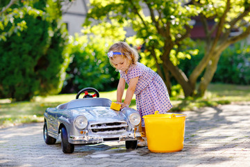 Cute gorgeous toddler girl washing big old toy car in summer garden, outdoors. Happy healthy little...