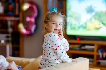 Cute little toddler girl in nightwear pajamas watching cartoons or movie on tv. Happy healthy baby child at home.