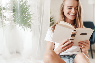 Image of laughing young woman reading book while sitting on bed