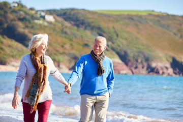 Loving Senior Couple Holding Hands As They Walk Along Shoreline Of Beach By Waves