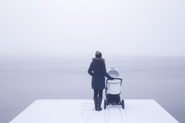 Young mother standing with baby stroller on snow covered footbridge and staring at lake. Mist over...