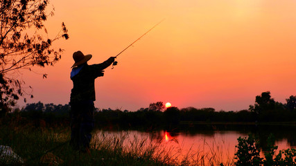 Silhouette fisherman with hat fishing on river bank and  blurred sunset sky background in evening time