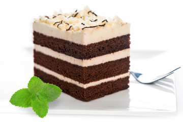 Delicious multilayer homemade cocoa sponge cake with sugar mass on a plate with a fork and mint leaf in close-up