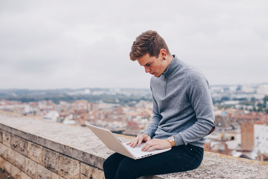 Blond young man sitting on a wall and typing on his laptop in Lleida