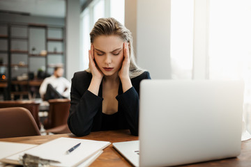 A beautiful blonde businesswoman sits in a loft office at a table with a laptop and suffers from headaches and work fatigue