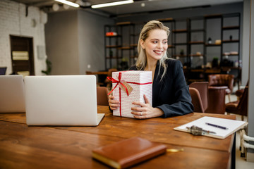A beautiful blonde businesswoman sits at a Desk in the office and holds a gift that was given by colleagues at work